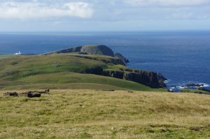 Colour photograph taken from the beacon site in Fair Isle. The view shows rough grassland sloping down from a high point towards a headland with the blue sea beyond, stretching off into a cloudy horizon.