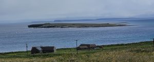 Colour photograph of the Holm of Papa (a small island) off Papa Westray. The photograph is taken from Papa Westray, looking across fields and houses into the bay in which the Holm lies.