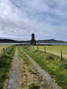 Colour photo of a stone church on a lightly cloudy day. It has a distinctive round tower. The church sits in the middle ground with an unmettled lane leading to it. The background is sea with hills beyond.