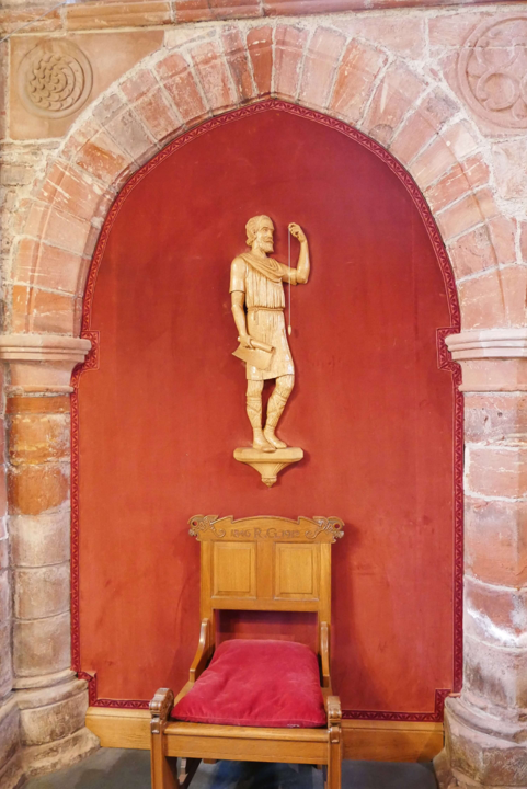 Colour photograph of the Kolr statuette in St. Magnus Cathedral, Kirkwall. The statuette depicts a man holding a rolled up plan in one hand and a plumb line in the other.