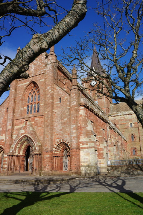 Colour photograph of St Magnus Cathedral, Kirkwall. The cathedral is made of a warm red sandstone and the photo is taken on a clear day with a deep blue sky.