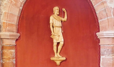 Colour photograph of the Kolr statuette in St. Magnus Cathedral, Kirkwall. The statuette depicts a man holding a rolled up plan in one hand and a plumb line in the other.