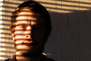 Man with striking eye, serious expression, sunlight coming through blinds