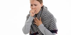 A woman coughing (possibly due to covid). She is wearing a grey sweater and has a shawl wrapped around her.