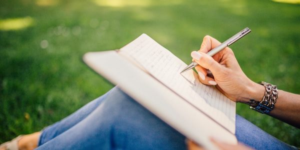 A person sat on the grass with a notebook on their lap, writing.