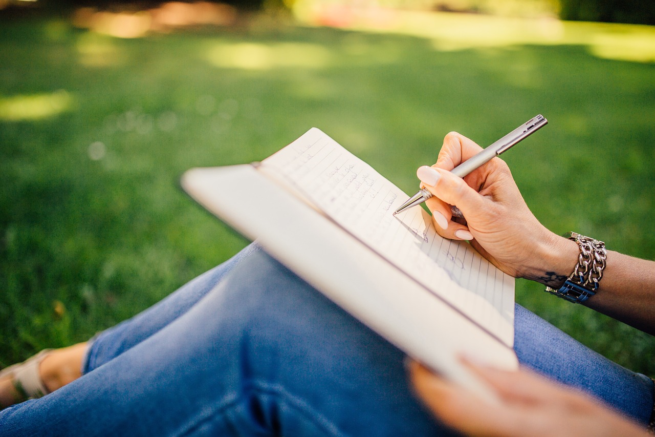 A person sat on the grass with a notebook on their lap, writing.