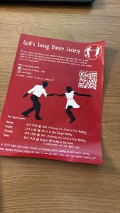 a flyer advertising the University of Nottingham's swing dance society. the poster is eye-catching. it has a crimson background. it lists a few upcoming meeting times. there is a large graphic of two black figures with no facial features, a man and a woman. the man has a white shirt and the woman has a white dress. the man and the woman are slightly far apart, but they are connected as they are holding hands, just with outstretched arms because they are dancing.