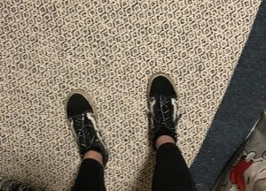 A photo taken from waist-height of a person's legs and feet as they stand on a flatweave rug with their shoes on