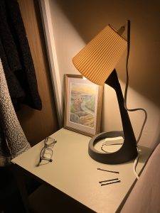 a small lamp atop a green metal bedside table. the lamp is switched on and casts yellow light onto the table and the wall