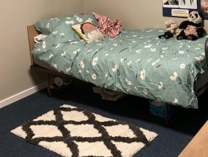 a bed in a student bedroom, with a pretty blue duvet cover with white daisies on. a fluffy black and white rug with a criss-cross pattern (white background, black criss-crosses) is on the floor below the bed, and it is nearly as long as the bed