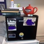 a black mini-fridge on top of a white mini-fridge, in a student room. the black fridge has two fridge magnets on it, and two pictures of the person's friends. the black fridge is being used as a bedside table. there is a bright red teapot on top of it, a pile of books, and a small basket containing a box of pens and a can of deodorant