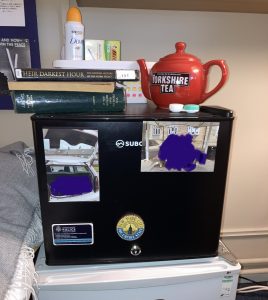 a black mini-fridge in a student room. the black fridge has two fridge magnets on it, and two pictures of the person's friends. the black fridge is being used as a bedside table. there is a bright red teapot on top of it, a pile of books, and a small basket containing a box of pens and a can of deodorant 