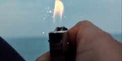 a hand holding a lighter against a blue background. the flame is lit and the blue sky and slightly darker blue sea are visible. the image is a bit dark, it looks like evening.