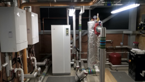 A room containing the Ground Source Heat Pump system, several white units can be seen with interconnected metal pipes. 