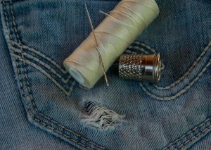 Photo of a pocket of jeans with a hole. There is also some thread, a needle and a thimble. 