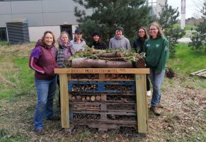 A group of people stand around a bug hotel smiling. The top of the bug hotel reads 'Insect Meditation Hotel'.
