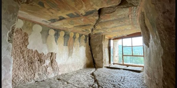 A cave with people shaped painting on the wall, which is crumbling away. A wooden window on the far wall.