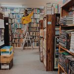 A researcher stands on a ladder in a library, surrounded by swathes of books piled on to bookshelves. The researcher reaches up to the top shelf to try and grab a specific book