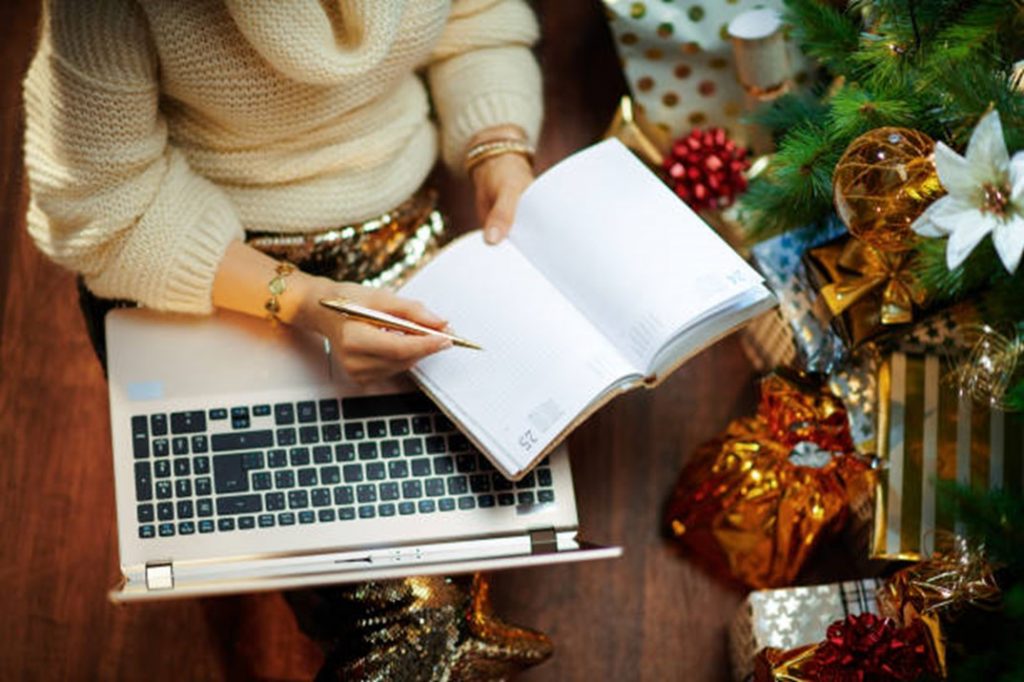 A student simultaneously writes in a notebook and holds a laptop, a christmas setting with presents and flowers is behind them