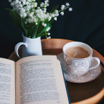 A book rests on a tray, alongside a cup of tea on a saucer, and a pot of flowers.