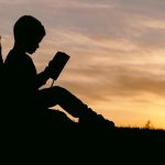 A figure of a young person sitting on the grass reading to themselves. The sun is setting in the background.