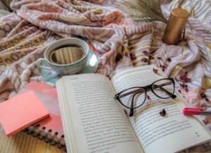 An arrangement of items including a book, glasses, a cup of coffee and a pink notebook, with black heart cut outs placed over the book