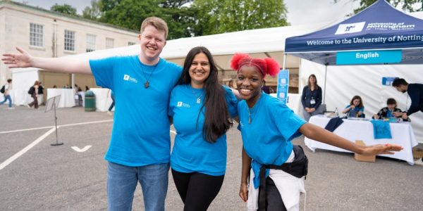 Three student ambassadors wearing the blue ambassador t-shirts, standing shoulder to shoulder with big smiles in front of the English tent at an Open Day