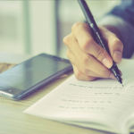 A person writing in a notebook with their phone on the side.